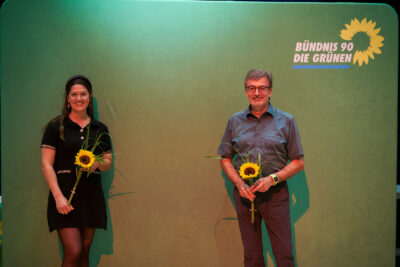 Das Bild zeigt Annika Hummel und Hans-Peter Behrens nach ihrer Nominierung vor einem grünen Hintergrund mit dem Parteilogo und Sonnenblumen in der Hand